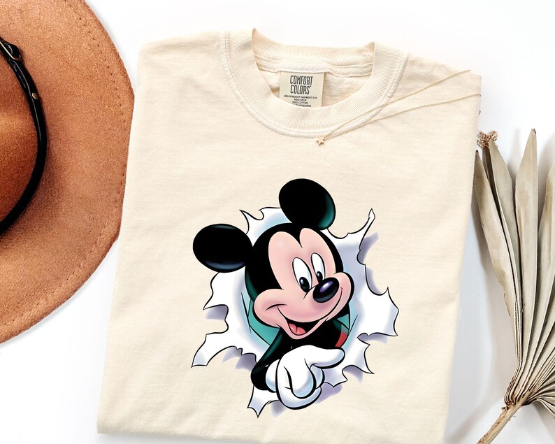 May include: A cream-colored t-shirt featuring a graphic of Mickey Mouse appearing to emerge from a torn paper effect. The character has black ears, a pink face, and white gloves. The shirt has a Comfort Colors brand label at the inner neckline.