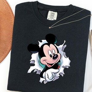 May include: A black crew neck t-shirt featuring a graphic of Mickey Mouse appearing to peek through a torn white paper effect. The shirt has a Comfort Colors brand label at the inner neckline and is made of cotton fabric with a short sleeve design.
