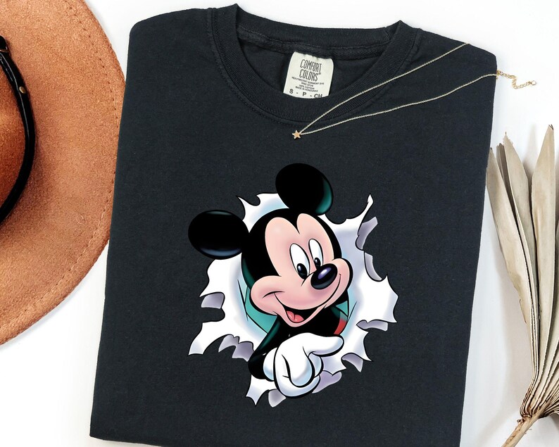 May include: A black crew neck t-shirt featuring a graphic of Mickey Mouse appearing to peek through a torn white paper effect. The shirt has a Comfort Colors brand label at the inner neckline and is made of cotton fabric with a short sleeve design.