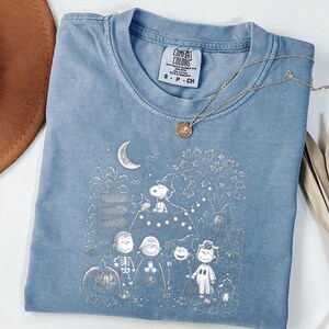 May include: A blue short-sleeved t-shirt featuring a white graphic print of Peanuts characters in Halloween costumes, including Snoopy in a witch hat, pumpkins, a crescent moon, and bats. The shirt has a Comfort Colors brand label at the neck.