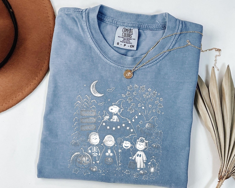 May include: A blue short-sleeved t-shirt featuring a white graphic print of Peanuts characters in Halloween costumes, including Snoopy in a witch hat, pumpkins, a crescent moon, and bats. The shirt has a Comfort Colors brand label at the neck.