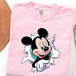 May include: A light pink crew neck t-shirt featuring a graphic of Mickey Mouse appearing to break through a white paper-like surface. The character has black ears, a tan face, and white gloves. The shirt has a Comfort Colors brand label at the neck.