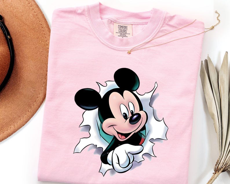 May include: A light pink crew neck t-shirt featuring a graphic of Mickey Mouse appearing to break through a white paper-like surface. The character has black ears, a tan face, and white gloves. The shirt has a Comfort Colors brand label at the neck.