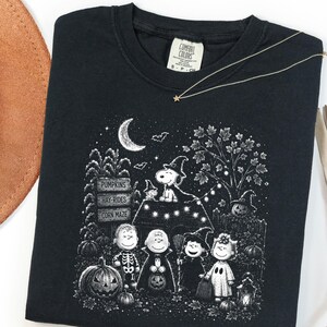 May include: A black crewneck t-shirt featuring a white graphic print of the Peanuts characters in Halloween costumes. The design includes Snoopy on a doghouse, pumpkins, a sign reading PUMPKINS, HAY-RIDES, CORN MAZE, and a crescent moon.
