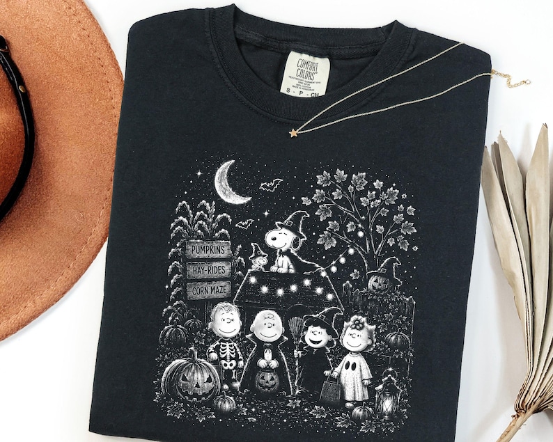 May include: A black crewneck t-shirt featuring a white graphic print of the Peanuts characters in Halloween costumes. The design includes Snoopy on a doghouse, pumpkins, a sign reading PUMPKINS, HAY-RIDES, CORN MAZE, and a crescent moon.