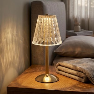 Peut inclure: Une petite lampe de table élégante avec un abat-jour clair et facetté et une base et une tige dorées. La lampe est allumée, émettant une lueur chaleureuse et invitante. La lampe est posée sur une table de chevet en bois.