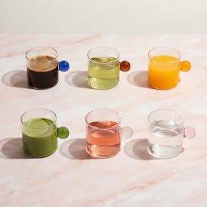 Könnte beinhalten: Ein Set aus sechs durchsichtigen Glasbechern, die jeweils einen kleinen, runden Griff aus farbigem Glas haben. Die Becher sind mit verschiedenen Flüssigkeiten gefüllt, darunter dunkler Kaffee, heller grüner Tee, Orangensaft, Matcha, rosa Flüssigkeit und klares Wasser.