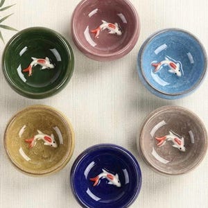 Peut inclure: Ensemble de sept petits bols en céramique de différentes couleurs, chacun orné d'une figurine de poisson koi blanc et rouge au fond. Les bols sont disposés en cercle, avec des couleurs telles que le vert, le rose, le bleu, le jaune, le marine et le gris.