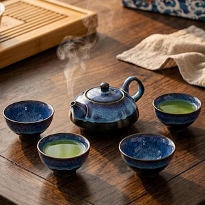 Peut inclure: Un service à thé en céramique émaillée bleu et noir. L'ensemble comprend une théière et quatre petites tasses remplies de thé vert. De la vapeur s'élève de la théière, suggérant du thé fraîchement infusé. L'ensemble est disposé sur une table en bois.