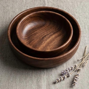 Peut inclure: Deux bols ronds en bois emboîtés avec une finition brun foncé et des motifs de grain de bois naturel visibles. Les bols sont lisses avec des bords incurvés et un intérieur profond, mettant en valeur la texture organique du bois.