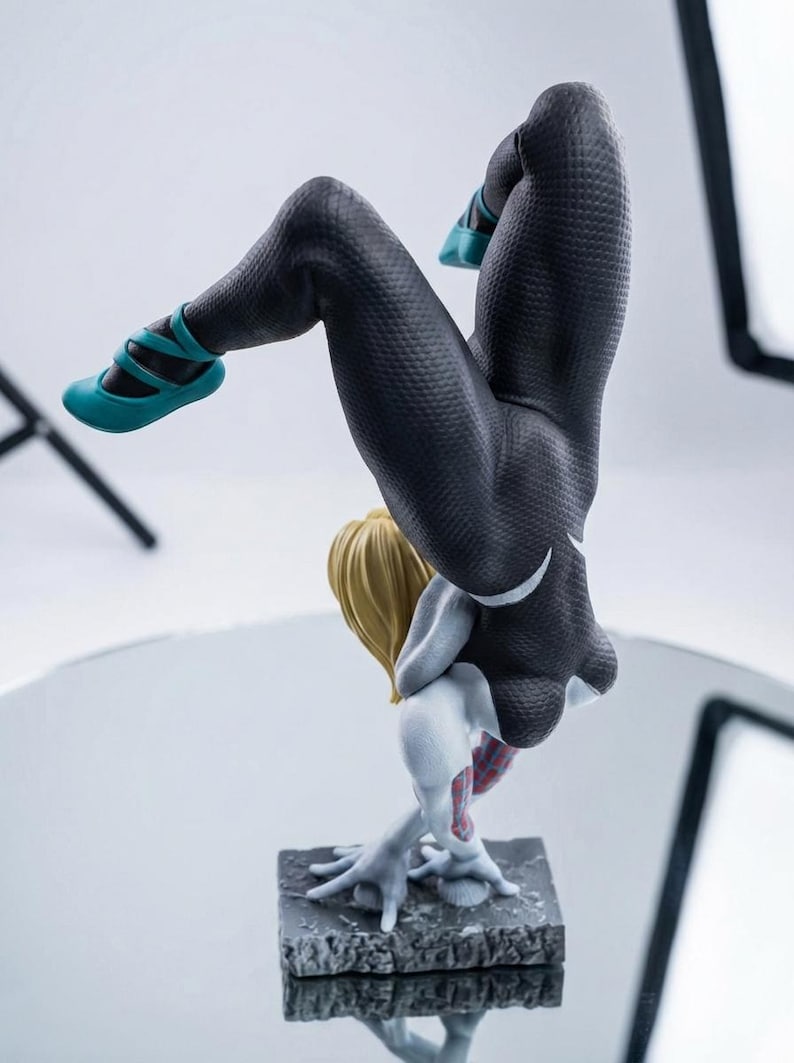 May include: A figurine of a character in a black and white suit with a spider-web pattern, blonde hair, and teal shoes. The figure is in a handstand pose on a textured gray base.