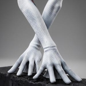 May include: A sculpture of pale, textured arms and hands, crossed at the wrists. The arms have a scale-like pattern, and the hands are open, resting on a dark, textured base. The background is a neutral gray.
