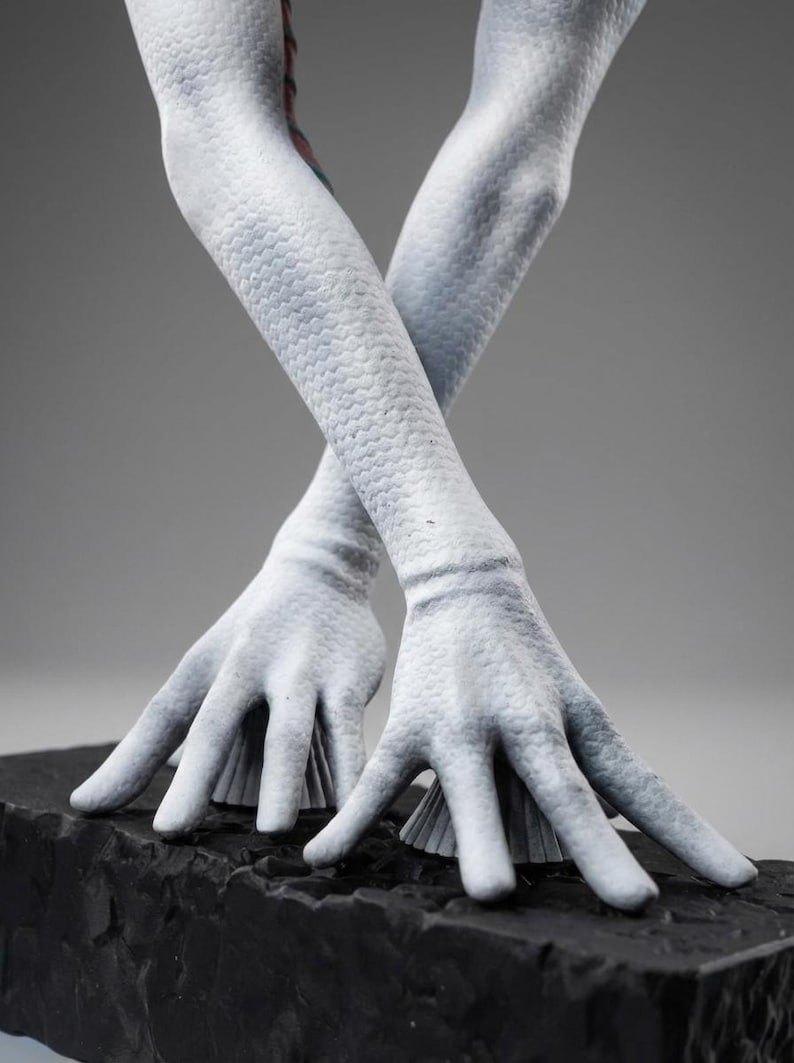 May include: A sculpture of pale, textured arms and hands, crossed at the wrists. The arms have a scale-like pattern, and the hands are open, resting on a dark, textured base. The background is a neutral gray.