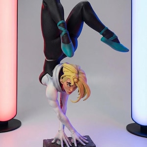 May include: A detailed figurine of a character in a dynamic handstand pose. The figure wears a black and white suit with teal ballet flats. The character has blonde hair and is posed on a rectangular base. The background has two colorful light columns.