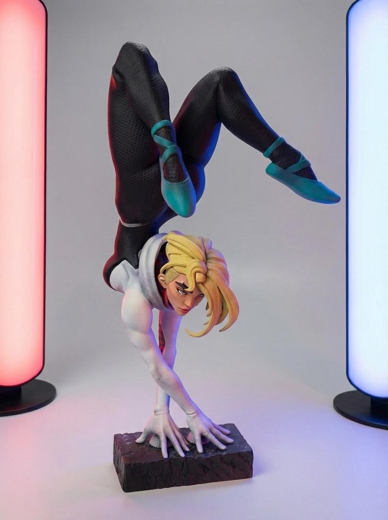 May include: A detailed figurine of a character in a dynamic handstand pose. The figure wears a black and white suit with teal ballet flats. The character has blonde hair and is posed on a rectangular base. The background has two colorful light columns.