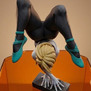 May include: A detailed figurine of a person in a handstand pose. The figure wears a textured black bodysuit and teal ballet flats. The figure's blonde hair is styled in a bun. The figure is supported by its hands on a stone base.
