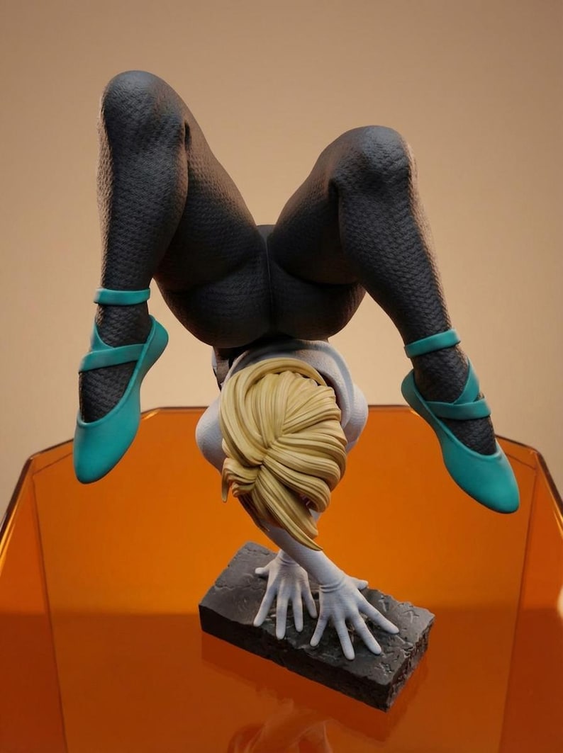 May include: A detailed figurine of a person in a handstand pose. The figure wears a textured black bodysuit and teal ballet flats. The figure's blonde hair is styled in a bun. The figure is supported by its hands on a stone base.