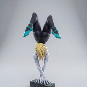 May include: A figurine of a person in a handstand, with blonde hair and a white and blue outfit. The figure's legs are clad in black leggings and turquoise shoes. The figure is balanced on a dark gray rectangular base.