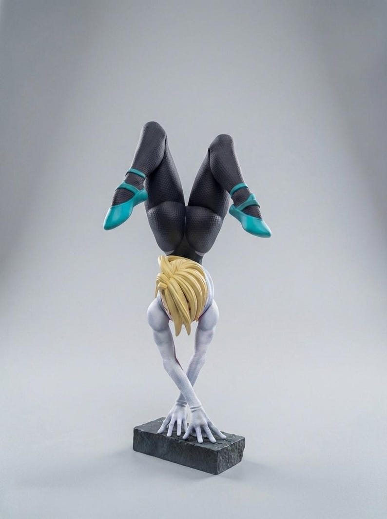 May include: A figurine of a person in a handstand, with blonde hair and a white and blue outfit. The figure's legs are clad in black leggings and turquoise shoes. The figure is balanced on a dark gray rectangular base.