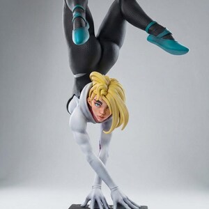 May include: A detailed figurine of a female superhero in a handstand pose. She wears a black and white suit with teal accents on her shoes. Her blonde hair is styled, and she is on a textured base.