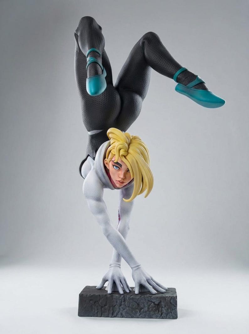 May include: A detailed figurine of a female superhero in a handstand pose. She wears a black and white suit with teal accents on her shoes. Her blonde hair is styled, and she is on a textured base.