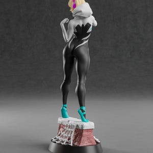 May include: A figurine of a female superhero in a black and white suit with a spiderweb design. She wears a white hooded jacket, teal high heels, and has blonde hair. The figurine stands on a red and white base.