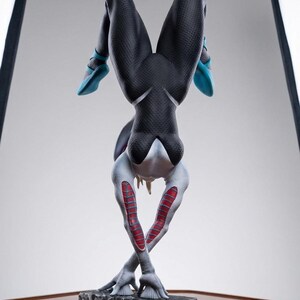 May include: A detailed statue of a character in a dynamic pose, upside down. The figure is primarily black with textured detailing, accented by light blue and red elements. It stands on a textured, dark gray base.