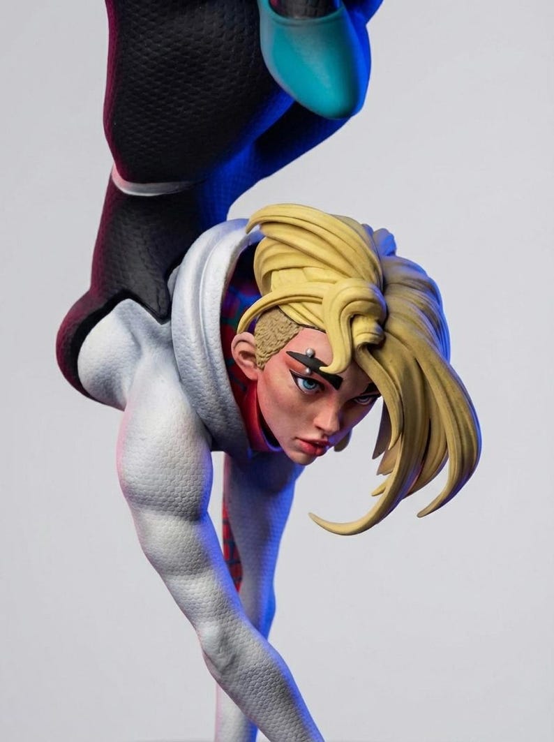 May include: A detailed figurine of a female superhero in a dynamic pose. She wears a white and black suit with a hood, and her blonde hair is styled in a unique fashion. The figure has blue eyes and a determined expression.