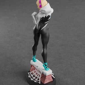 May include: A figurine of a female character in a black and white bodysuit, teal heels, and a white hooded jacket. The figure has blonde hair and pink headphones. The base features a brick-like structure with a white spiderweb design.