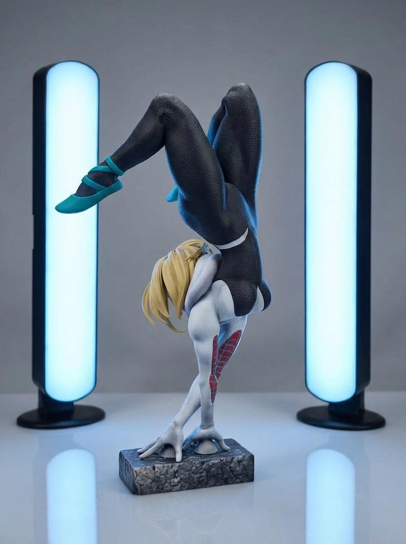 May include: A detailed figurine of a character in a black and white suit, with blonde hair, striking a handstand pose. The figure is on a textured base, with teal shoes. Two tall, glowing blue lights flank the figure.