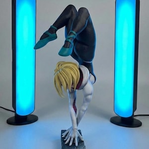 May include: A statue of a superheroine in a dynamic handstand pose, with blonde hair and a white and black suit. The figure is flanked by two tall, black, rectangular light fixtures emitting a bright blue glow.