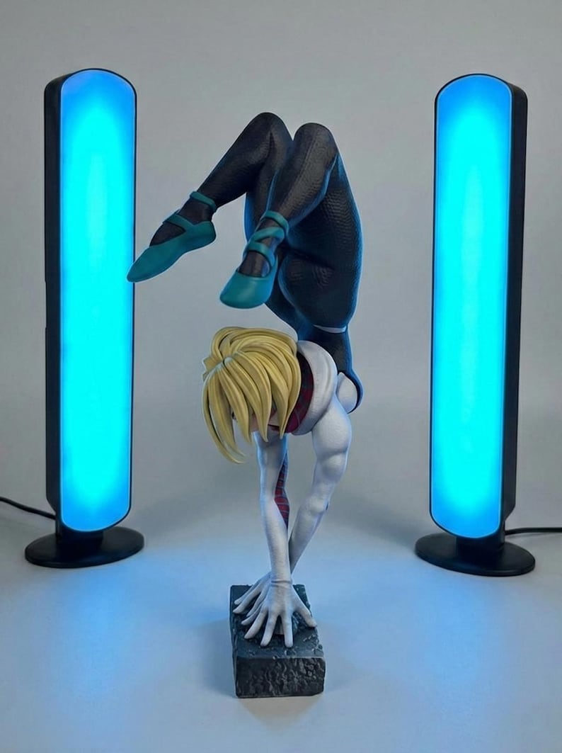 May include: A statue of a superheroine in a dynamic handstand pose, with blonde hair and a white and black suit. The figure is flanked by two tall, black, rectangular light fixtures emitting a bright blue glow.