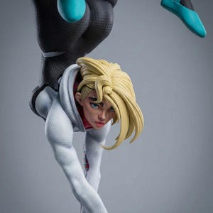 May include: A detailed figurine of a female character in a dynamic pose. She wears a white hoodie, a black bodysuit, and teal ballet flats. Her blonde hair is styled, and she has blue eyes. The figure is set against a gray backdrop.