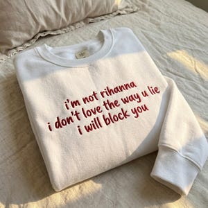 May include: White crew neck sweatshirt with the embroidered text "i'm not rihanna i don't love the way u lie i will block you" in red. The sweatshirt is folded on a bed.