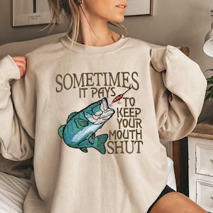 Könnte beinhalten: Beiges Sweatshirt mit dem Text "SOMETIMES IT PAYS TO KEEP YOUR MOUTH SHUT". Das Design zeigt einen blau-grünen Fisch mit einem Angelköder. Das Sweatshirt hat eine neutrale Farbe.