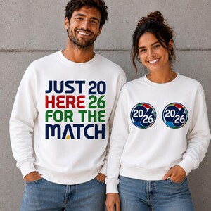 Puede incluir: Dos personas con sudaderas blancas. Una tiene el texto "JUST 20 HERE 26 FOR THE MATCH" en varios colores. La otra tiene dos gr&aacute;ficos de bal&oacute;n de f&uacute;tbol con "20/26" en su interior. Ambas son para un partido.
