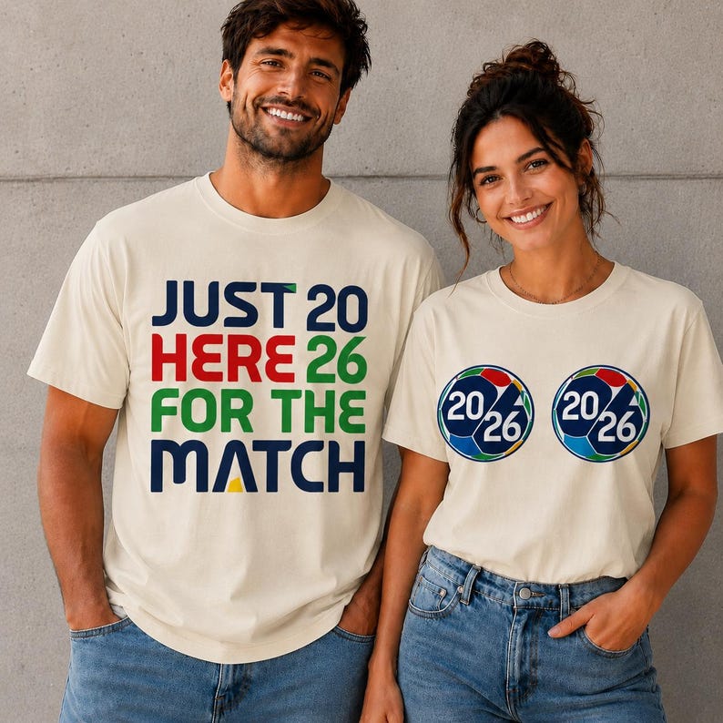 Puede incluir: Dos personas con camisetas color crema. Una camiseta dice "JUST 20 HERE 26 FOR THE MATCH" en azul, rojo y verde. La otra tiene dos dise&ntilde;os de bal&oacute;n de f&uacute;tbol con "20/26" en su interior.