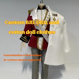 Puede incluir: Un conjunto de ropa para muñeco que incluye una chaqueta militar roja con puños negros y adornos dorados, pantalones grises y una capa blanca sobre un hombro. El atuendo presenta una banda blanca, medallas doradas y un cuello alto con perlas.