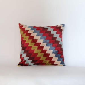 Op de afbeelding: Vierkant decoratief kussen met een geometrisch zigzagpatroon in rood, blauw, groen en crème. Het kussen heeft een zacht, getextureerd uiterlijk, waarschijnlijk van fluweel. Een decoratief item voor in huis.