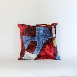 May include: Square cushion featuring a geometric design in red, blue, yellow, and grey. The cushion has a soft, velvety texture and a contemporary design, ideal for adding a splash of colour to any room. The cushion is square in shape.
