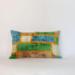 Op de afbeelding: Rechthoekig decoratief kussen met een geometrisch patroon. Het kussen heeft een patchwork ontwerp met blauwe, groene, bruine en crème secties, omlijnd met oranje stiksels. Een zacht, pluche woondecoratie-item.