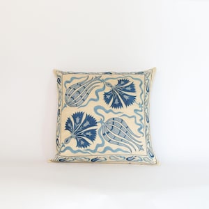 May include: Square decorative pillow with a cream-colored background and embroidered blue floral designs. The pillow features stylized flowers and wavy lines, with a border of small blue floral patterns.
