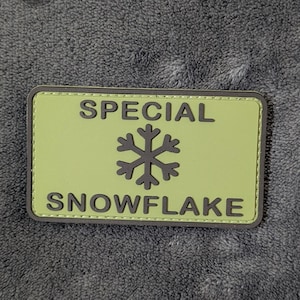 May include: A rectangular patch with a light green background and a dark gray border. The text "SPECIAL" is printed in dark gray above a dark gray snowflake icon, with the word "SNOWFLAKE" printed in dark gray below the icon.
