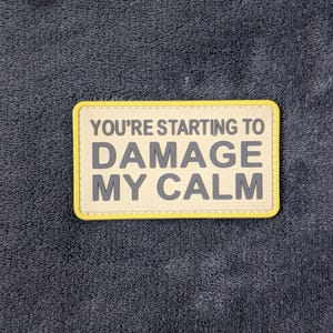 Op de afbeelding: Een rechthoekige patch met de tekst "YOU'RE STARTING TO DAMAGE MY CALM" in grijze letters. De patch heeft een gele rand en een lichtbeige achtergrond. De patch ligt op een donkergrijze, getextureerde ondergrond.