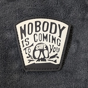 May include: A black and white patch with the text "NOBODY IS COMING TO SAVE YOU" in bold letters. The design includes a skull and crossbones graphic. The patch has a black border and is set against a dark gray textured background.