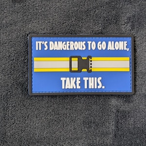 May include: A rectangular patch with a blue background and black border. The patch features the text "IT'S DANGEROUS TO GO ALONE, TAKE THIS." in white. A yellow and gray design resembling a belt buckle is in the center.