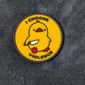 Può includere: Una toppa rotonda gialla con un'anatra dei cartoni animati che tiene un coltello. L'anatra ha un becco e zampe rosse, e il testo "I CHOOSE VIOLENCE" è scritto intorno all'anatra in lettere nere. La toppa ha un bordo nero.