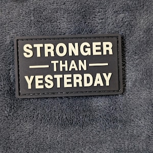 May include: A rectangular black patch with raised off-white lettering that reads STRONGER THAN YESTERDAY. The patch features a clean, bold sans-serif font with a horizontal line separating the words, designed for attachment to fabric surfaces.