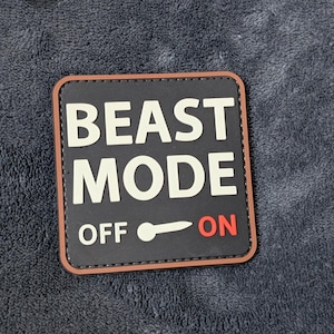 May include: A square patch with the words "BEAST MODE" in white block letters on a black background. The word "OFF" is on the left, and "ON" is in red on the right, with a white switch graphic in between. The patch has a brown border.