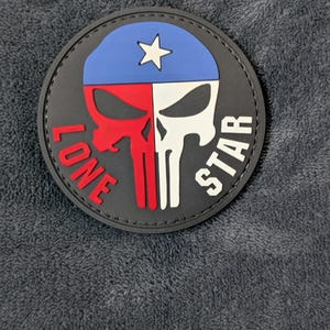 May include: A circular black patch featuring a stylized skull graphic with a blue top section containing a white star. The left side of the skull is red and the right side is white. The text LONE is in red on the left and STAR is in white on the right.